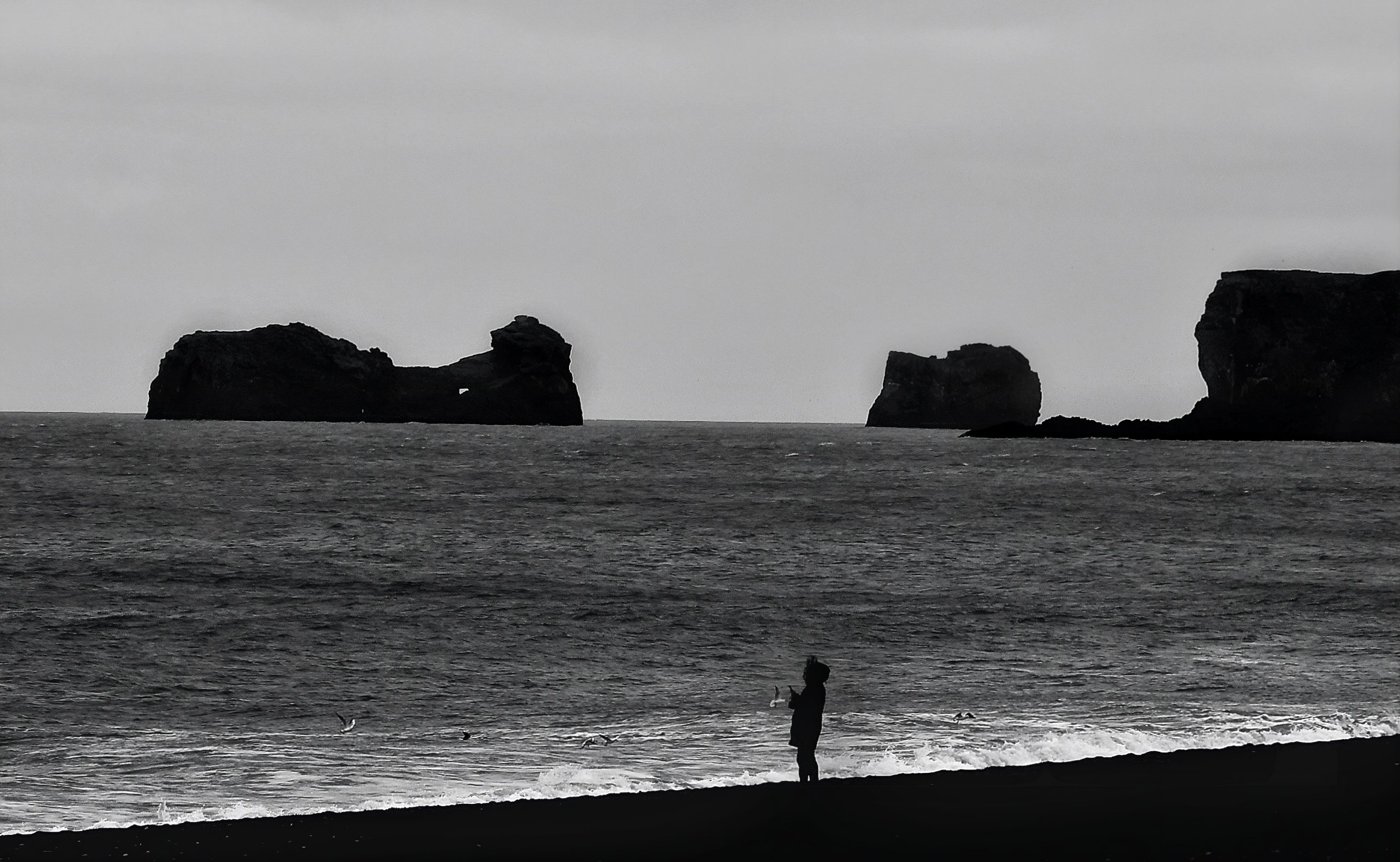 Black-and-white photograph by Rebecca Kavalchuck: a lone figure on a dark beach facing sea stacks and gulls